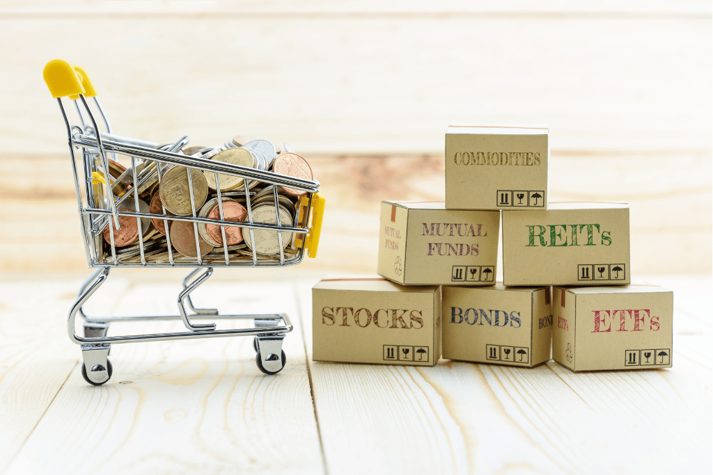 Shopping cart with coins next to s stack of boxes with "commodities", "mutual funds", "REITs", "stocks", "bonds", and "ETFs" written on them. Denoting diversifying in different investment assets.